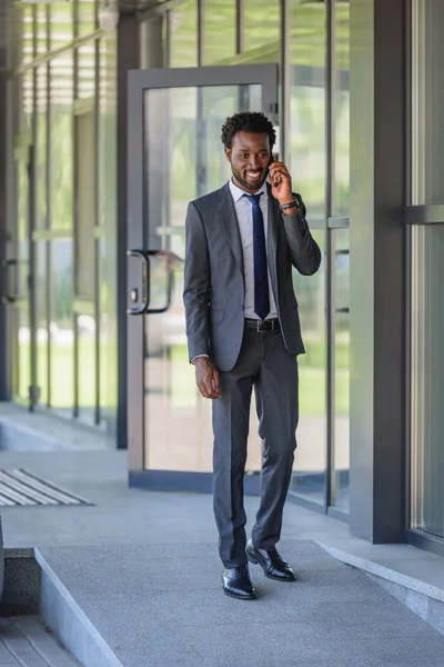 Sorridente empresário afro-americano falando no smartphone enquanto caminha ao longo do escritório buiding — Fotografia de Stock