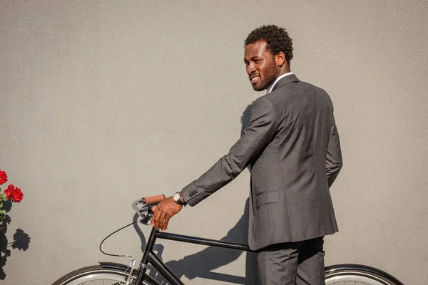 Bel homme d'affaires afro-américain en costume debout au mur avec vélo — Photo de stock