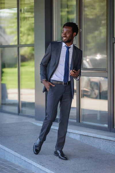 Alegre hombre de negocios afroamericano con teléfono inteligente caminando a lo largo del edificio de oficinas y cogido de la mano en el bolsillo - foto de stock