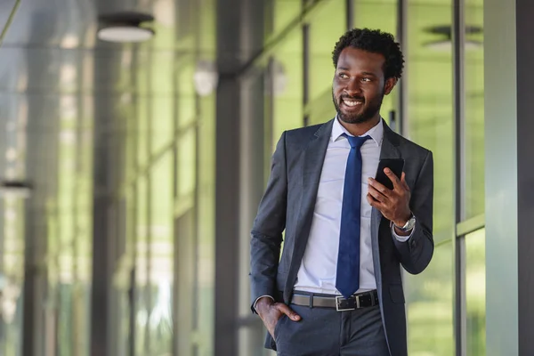 Homme d'affaires afro-américain souriant avec smartphone marchant près de l'immeuble de bureaux et tenant la main dans la poche — Photo de stock