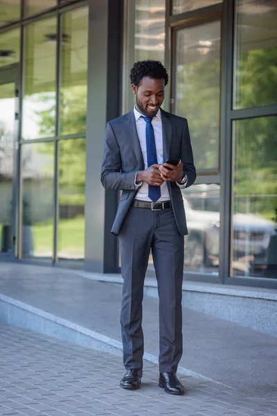 Positiv afrikanisch-amerikanischer Geschäftsmann mit Smartphone zu Fuß in der Nähe von Bürogebäude — Stockfoto