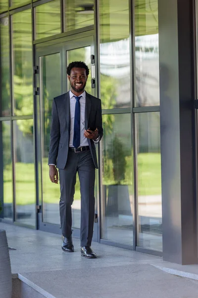 Lächelnder afrikanisch-amerikanischer Geschäftsmann mit Smartphone in der Nähe von Bürogebäuden — Stockfoto