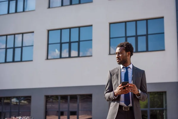 Selbstbewusster afrikanisch-amerikanischer Geschäftsmann schaut weg, während er sein Smartphone in der Hand hält — Stockfoto