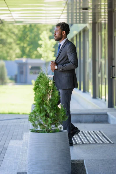 Hombre de negocios afroamericano confiado mirando hacia otro lado mientras que está parado cerca del edificio de oficinas - foto de stock