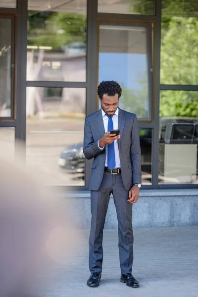 Enfoque selectivo del hombre de negocios afroamericano utilizando el teléfono inteligente mientras está cerca del edificio de oficinas - foto de stock