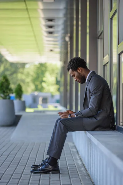 Riflessivo uomo d'affari afroamericano americano utilizzando smartphone mentre seduto sul parapetto vicino all'edificio degli uffici — Foto stock