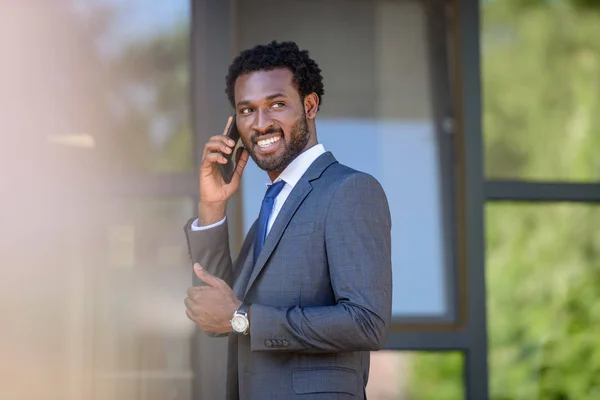Enfoque selectivo de sonreír hombre de negocios afroamericano hablando en el teléfono inteligente cerca del edificio de oficinas - foto de stock