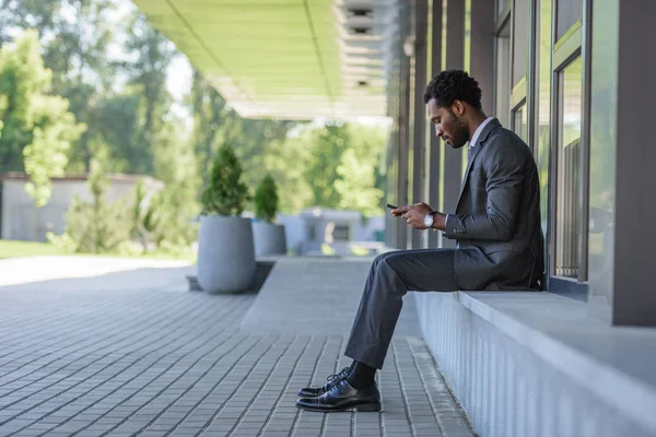 Uomo d'affari afroamericano premuroso utilizzando smartphone mentre seduto sul parapetto — Foto stock