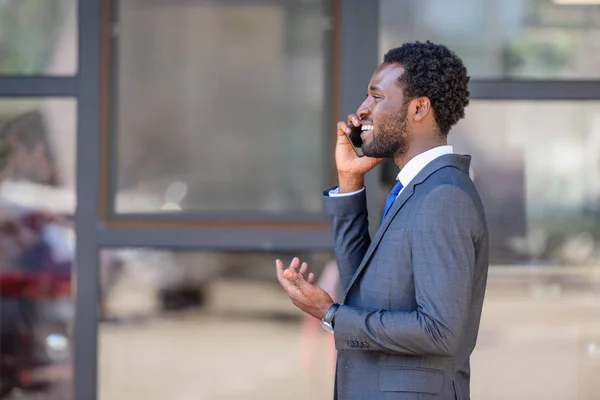 Alegre hombre de negocios afroamericano hablando en el teléfono inteligente cerca del edificio de oficinas - foto de stock