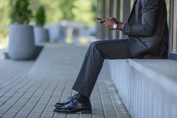 Vista recortada del hombre de negocios afroamericano utilizando el teléfono inteligente mientras está sentado en parapeto - foto de stock