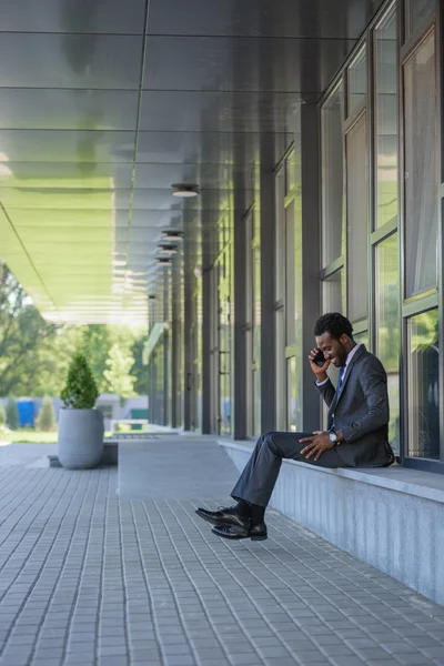 Улыбающийся африканский американский бизнесмен разговаривает по смартфону, сидя возле офисного здания — стоковое фото