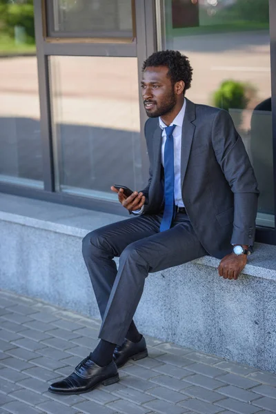 Pensativo hombre de negocios afroamericano sosteniendo teléfono inteligente mientras está sentado en parapeto - foto de stock