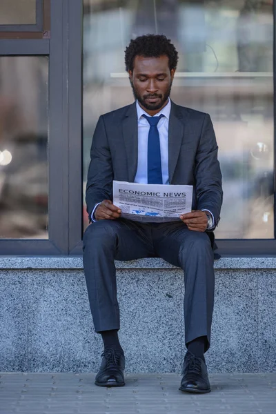 Attentive african american businessman reading newspaper while sitting near office building — Stock Photo
