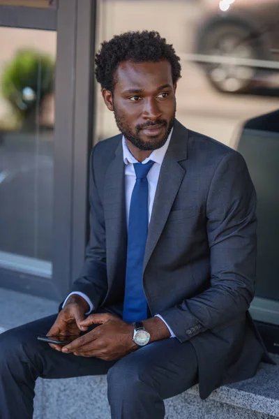 Nachdenklicher afrikanisch-amerikanischer Geschäftsmann hält Smartphone in der Hand und schaut weg, während er auf Brüstung sitzt — Stockfoto