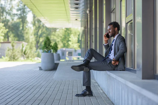 Allegro uomo d'affari afroamericano che parla su smartphone e tiene la mano sull'anca mentre si siede vicino all'edificio degli uffici — Foto stock