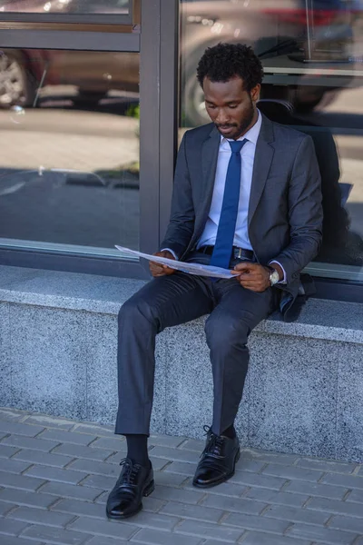 Focalizzato afroamericano uomo d'affari lettura giornale mentre seduto vicino edificio per uffici — Foto stock
