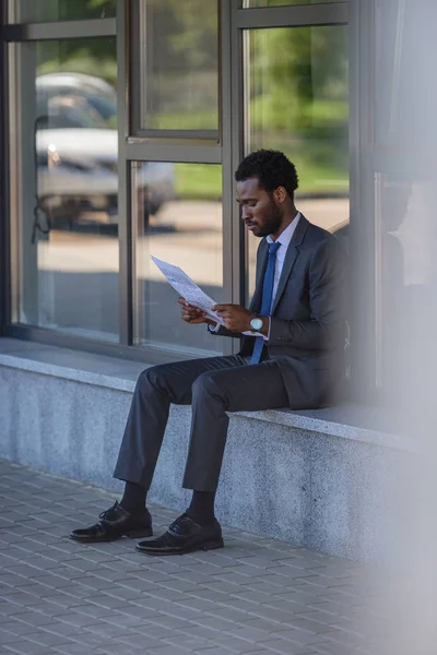 Foco selectivo del empresario afroamericano atento que lee el periódico mientras que está sentado cerca del edificio de oficinas - foto de stock