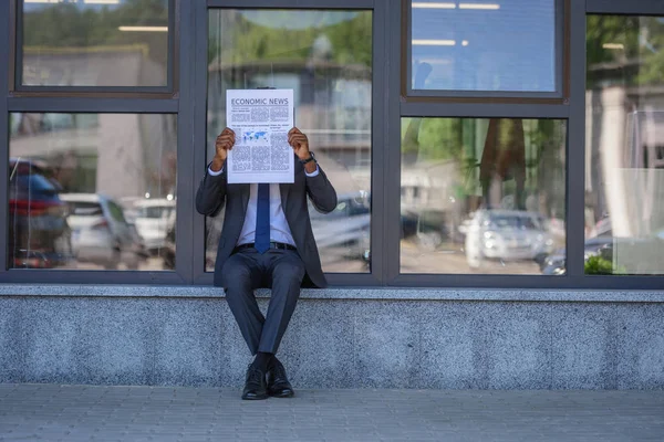 African american businessman reading economic news newspaper while sitting near office building with glass facade — Stock Photo