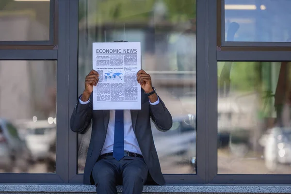 Uomo d'affari africano americano che legge il giornale di notizie economiche mentre si siede vicino all'edificio degli uffici con facciata in vetro — Foto stock