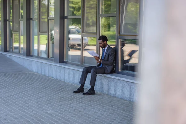 Selective focus of concentrated african american businessman reading newspaper while sitting on parapet of office building — Stock Photo