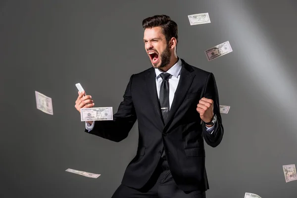 Збуджений бізнесмен зі смартфоном поблизу падаючих доларових банкнот на сірому — стокове фото