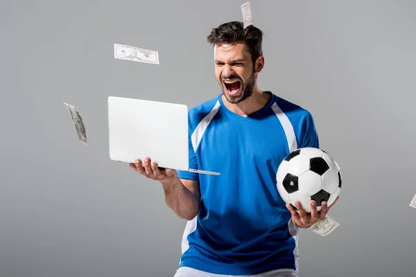 Jugador de fútbol emocionado con la pelota y el ordenador portátil cerca de caer dinero en gris - foto de stock
