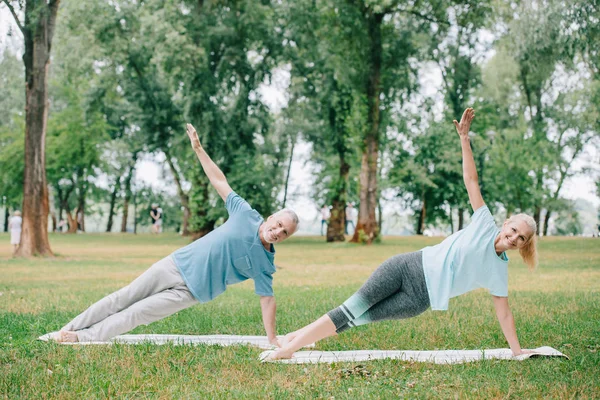 Hombre y mujer positivos y maduros haciendo ejercicio de tablón lateral mientras practican yoga en el césped en el parque - foto de stock