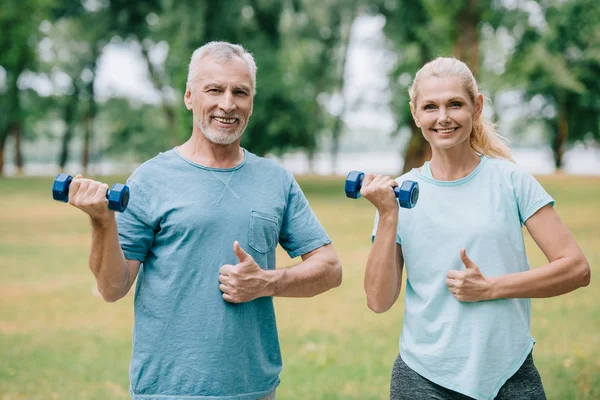 Smiling mature sportsman and sportswoman holding barbells and showing thumbs up while smiling at camera — Stock Photo