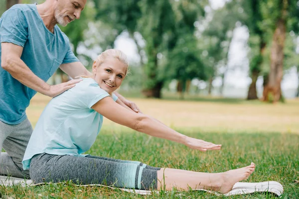 Guapo maduro hombre ayudando sonriente mujer practicando yoga en parque - foto de stock