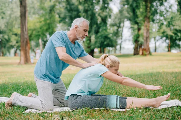 Красивый зрелый мужчина помогает женщине практиковать позу йоги на газоне в парке — стоковое фото
