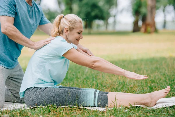 Vista parcial del hombre ayudando a la mujer sonriente a practicar yoga en el césped en el parque - foto de stock
