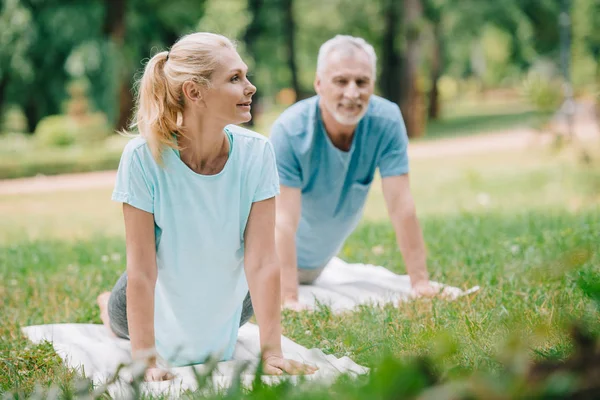 Селективный фокус улыбающихся, зрелых мужчин и женщин, практикующих йогу на ковриках для йоги в парке — стоковое фото
