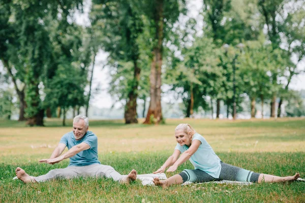 Sonriente, hombre y mujer maduros practicando yoga sobre esteras en parque - foto de stock