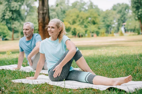 Sorrindo homem maduro e mulher praticando ioga enquanto sentado em tapetes de ioga no parque — Fotografia de Stock