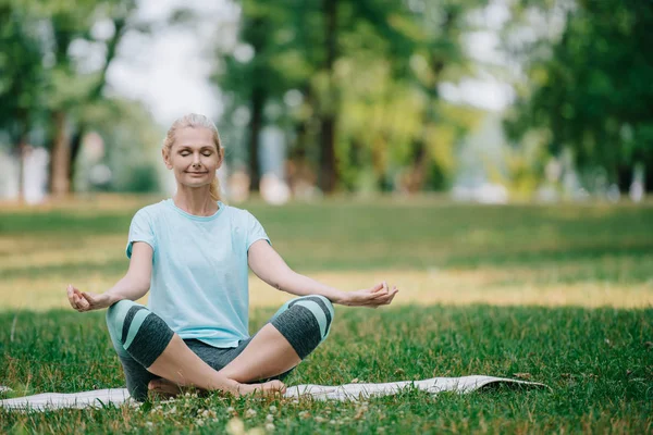 Bella donna matura meditando con gli occhi chiusi mentre seduto in posa di loto sul prato nel parco — Foto stock