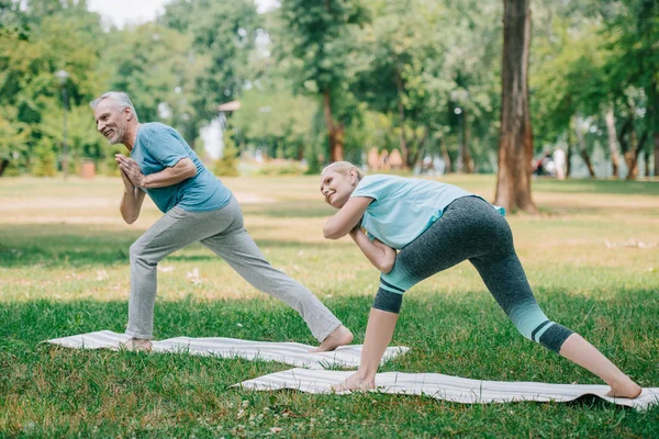 Hombre y mujer maduros practicando yoga mientras están de pie sobre esteras de yoga en el parque - foto de stock