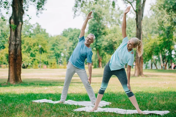 Hombre y mujer sonrientes y maduros de pie en poses guerreras mientras practican yoga en el parque - foto de stock