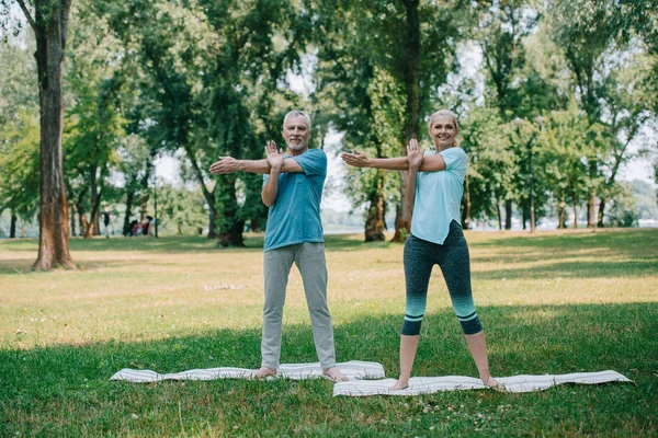 Hombre y mujer positivos y maduros practicando yoga mientras están de pie en las esteras de yoga en el parque - foto de stock