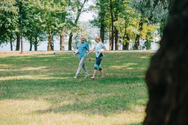 Foco seletivo de homem maduro e mulher segurando tapetes de ioga enquanto caminham juntos no parque — Fotografia de Stock