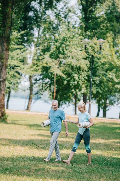 Cheerful mature man and woman walking together in park while holding yoga mats — Stock Photo