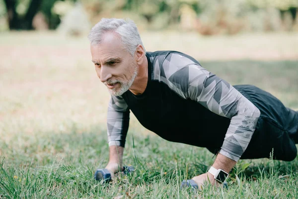 Positive mature sportsman doing push ups with barbells on lawn in park — Stock Photo