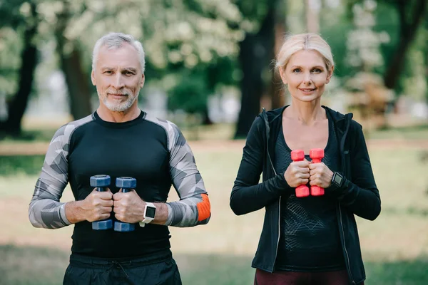 Positive, mature sportsman and sportswoman holding barbells and smiling at camera — Stock Photo