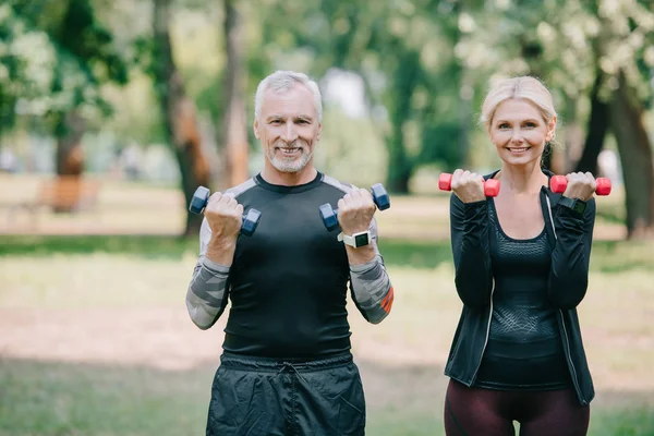 Cheerful, mature sportsman and sportswoman smiling at camera while training with barbells — Stock Photo