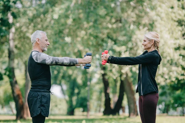 Side view of smiling mature sportsman and sportswoman training with barbells in park and looking at each other — Stock Photo