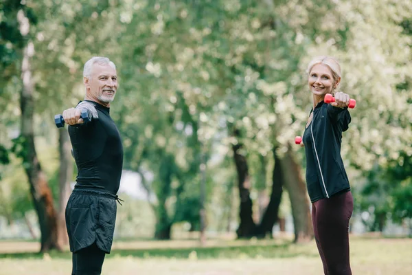 Cheerful, mature sportsman and sportswoman training with dumbbells in park and smiling at camera — Stock Photo