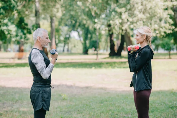 Side view of mature sportsman and sportswoman training with barbells in park while looking at each other — Stock Photo