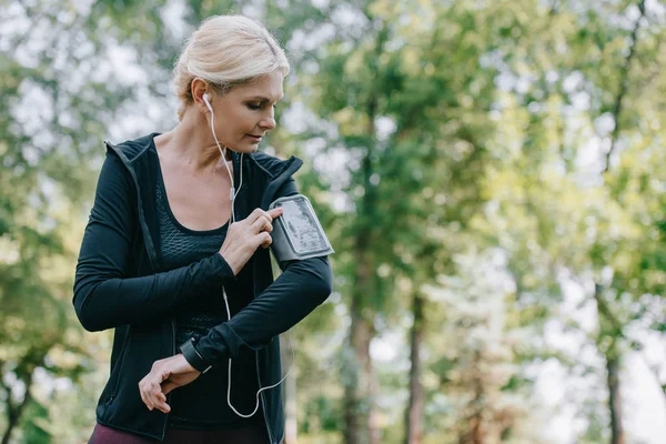 Attractive, mature sportswoman listening music in earphones in park — Stock Photo