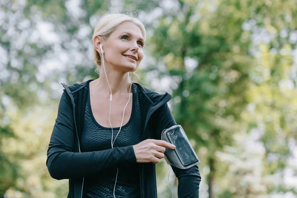 Smiling mature sportswoman looking away while listening music in earphones — Stock Photo