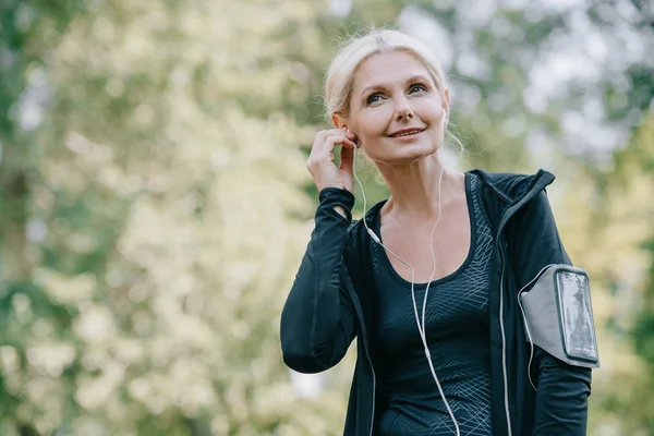 Smiling mature sportswoman looking away while listening music in earphones — Stock Photo