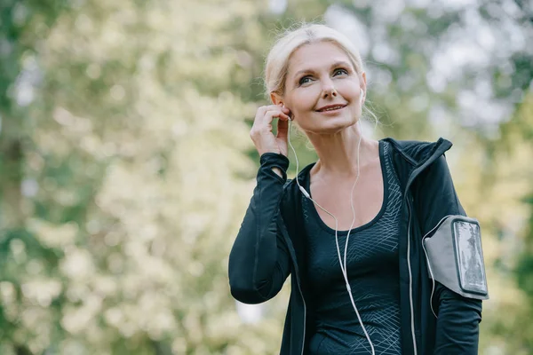 Lächelnde reife Sportlerin, die wegschaut, während sie Musik im Kopfhörer hört — Stockfoto